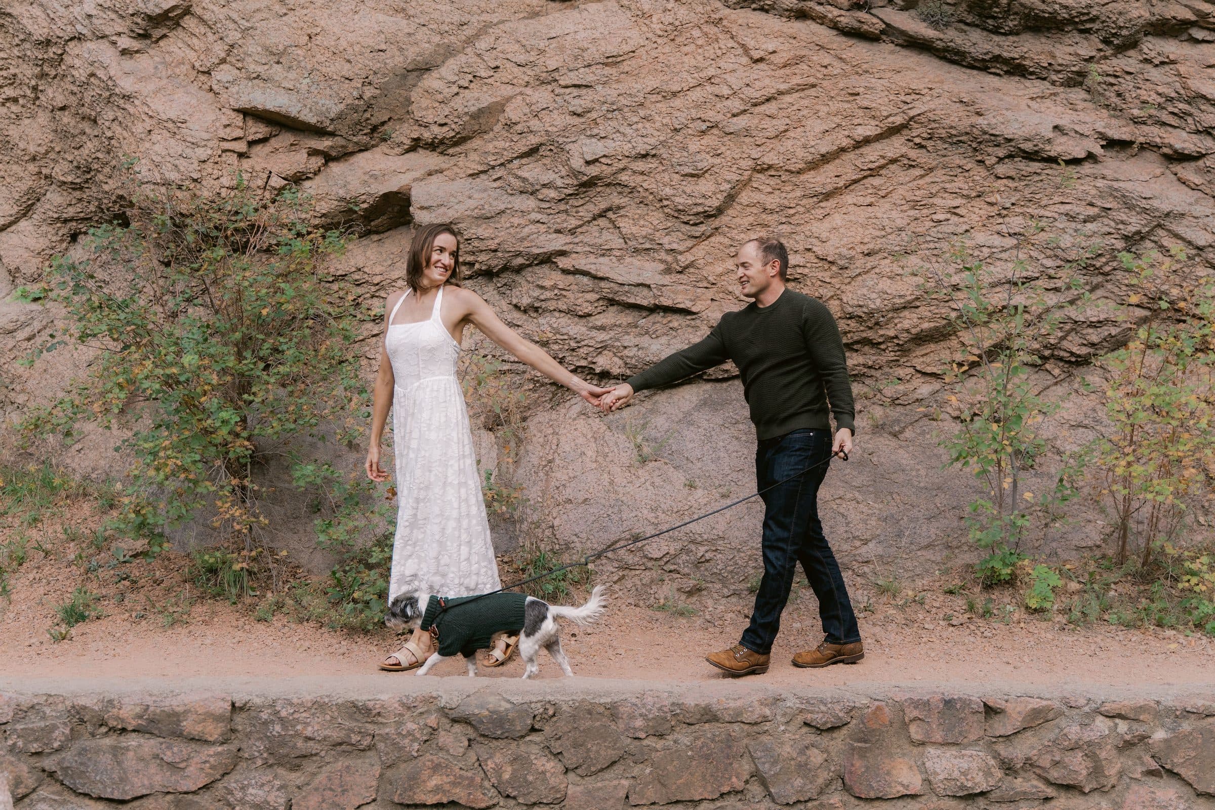 Couple holding hands and walking their dog along a red rock wall