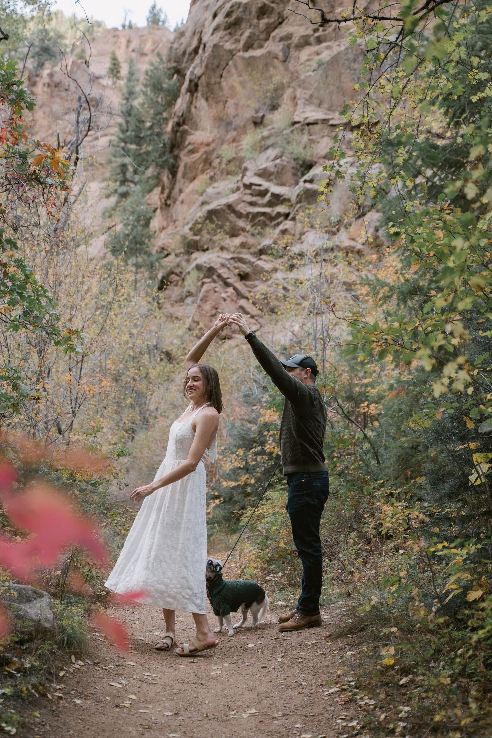 Man twirling his partner on a canyon trail with their dog beside them