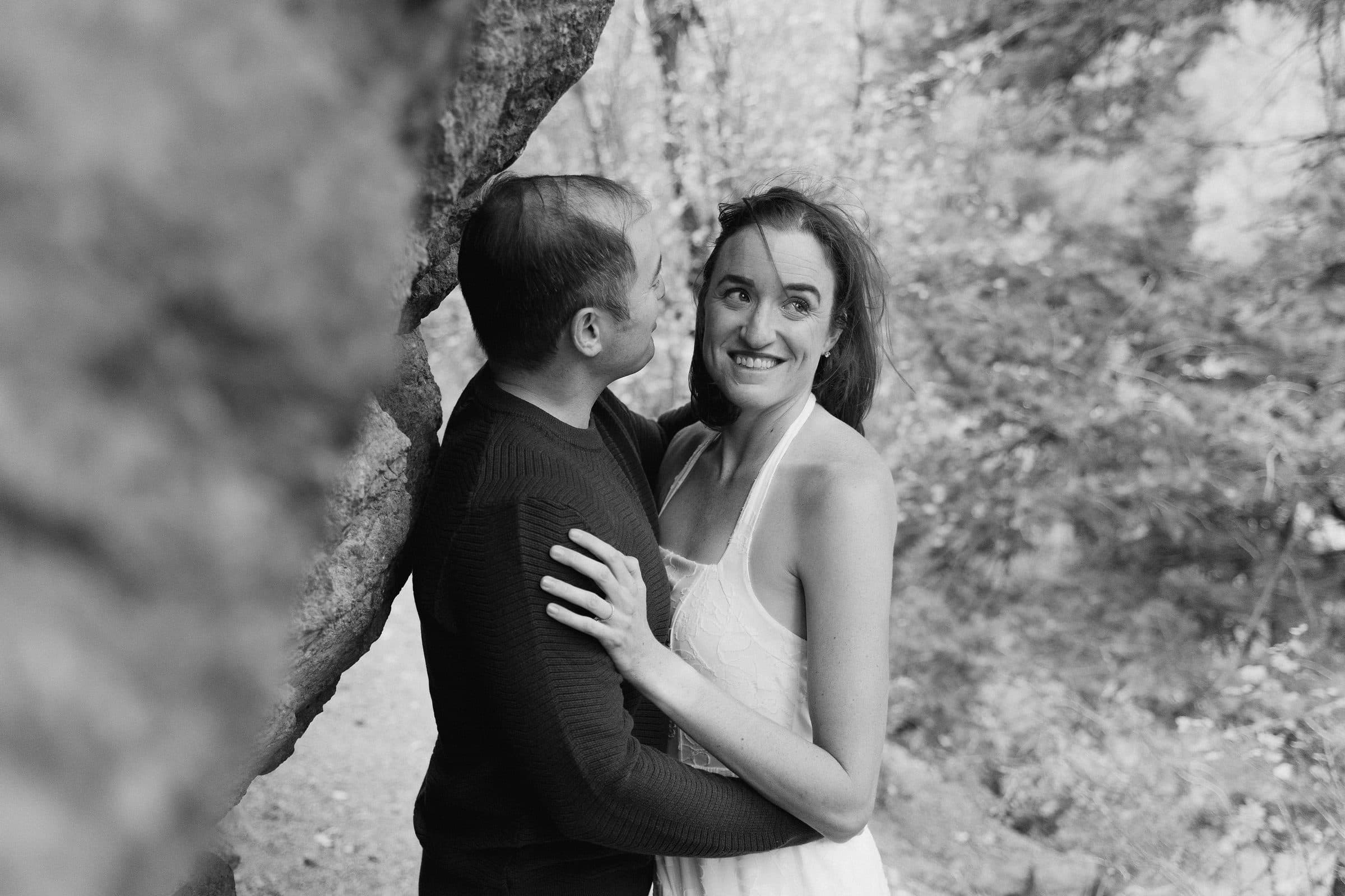 Black and white of a couple embracing beside a boulder, smiling at each other