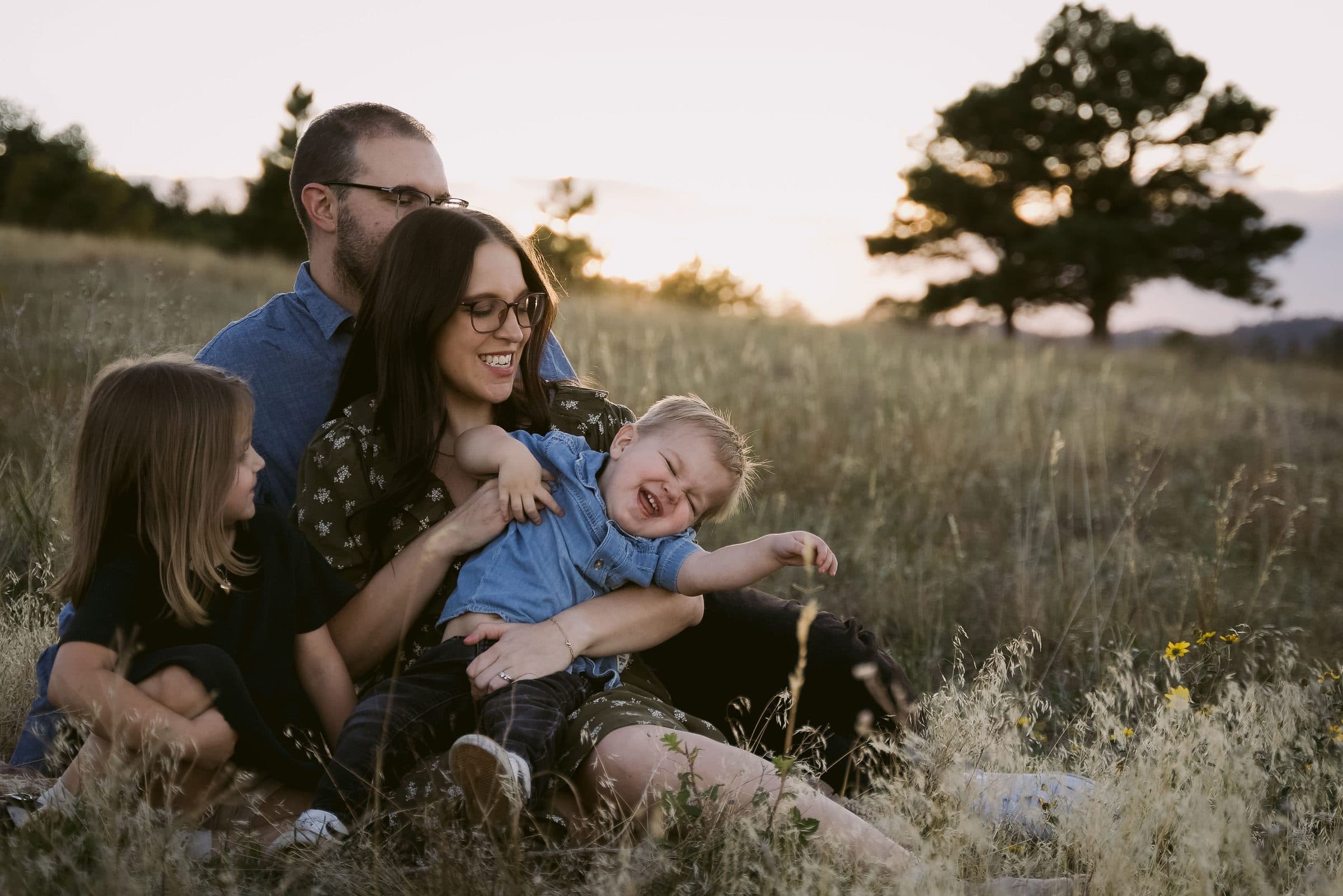 Family of four laughing together in the grass as the toddler wriggles