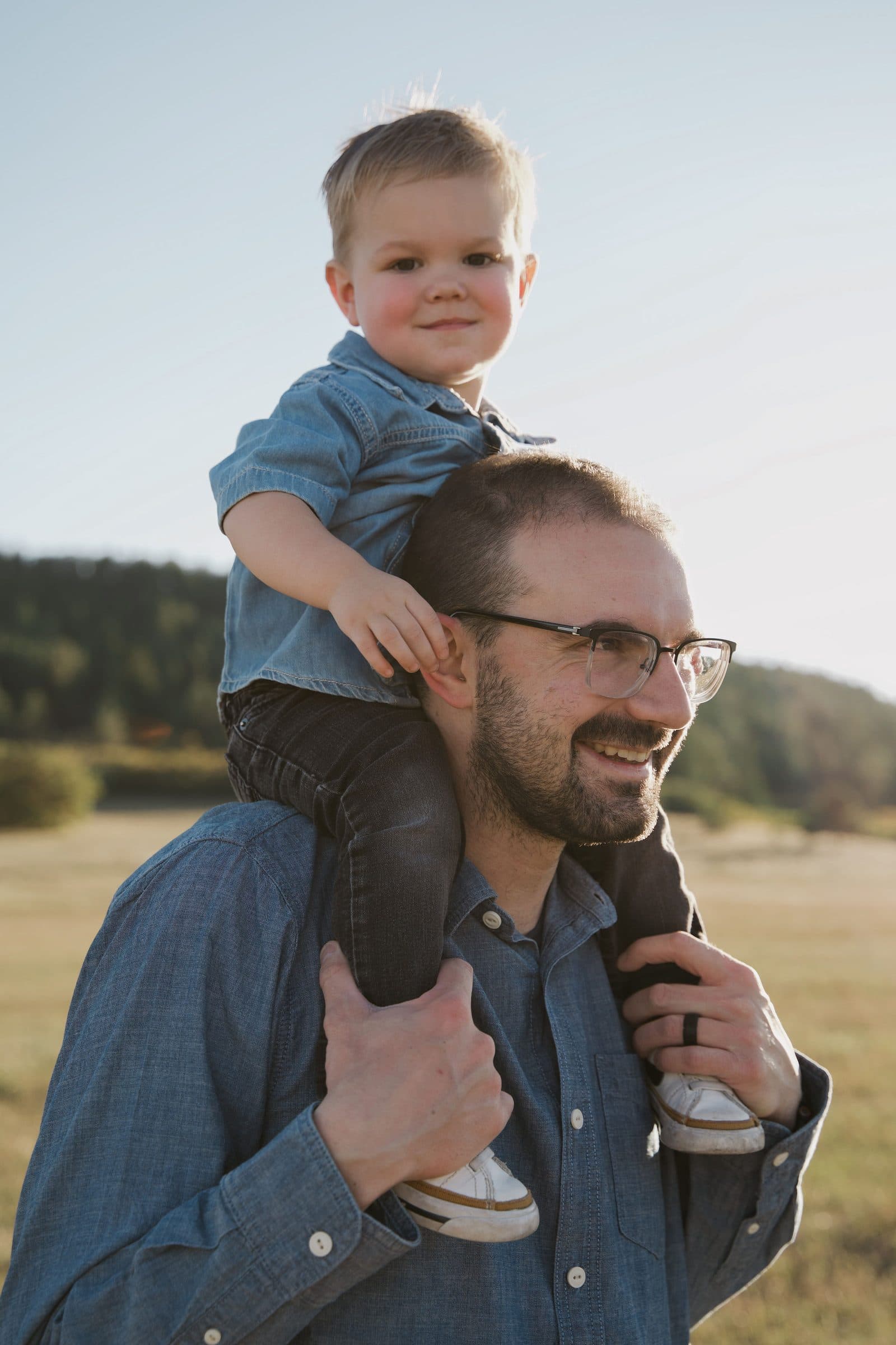 Toddler riding on his smiling father's shoulders in an open field