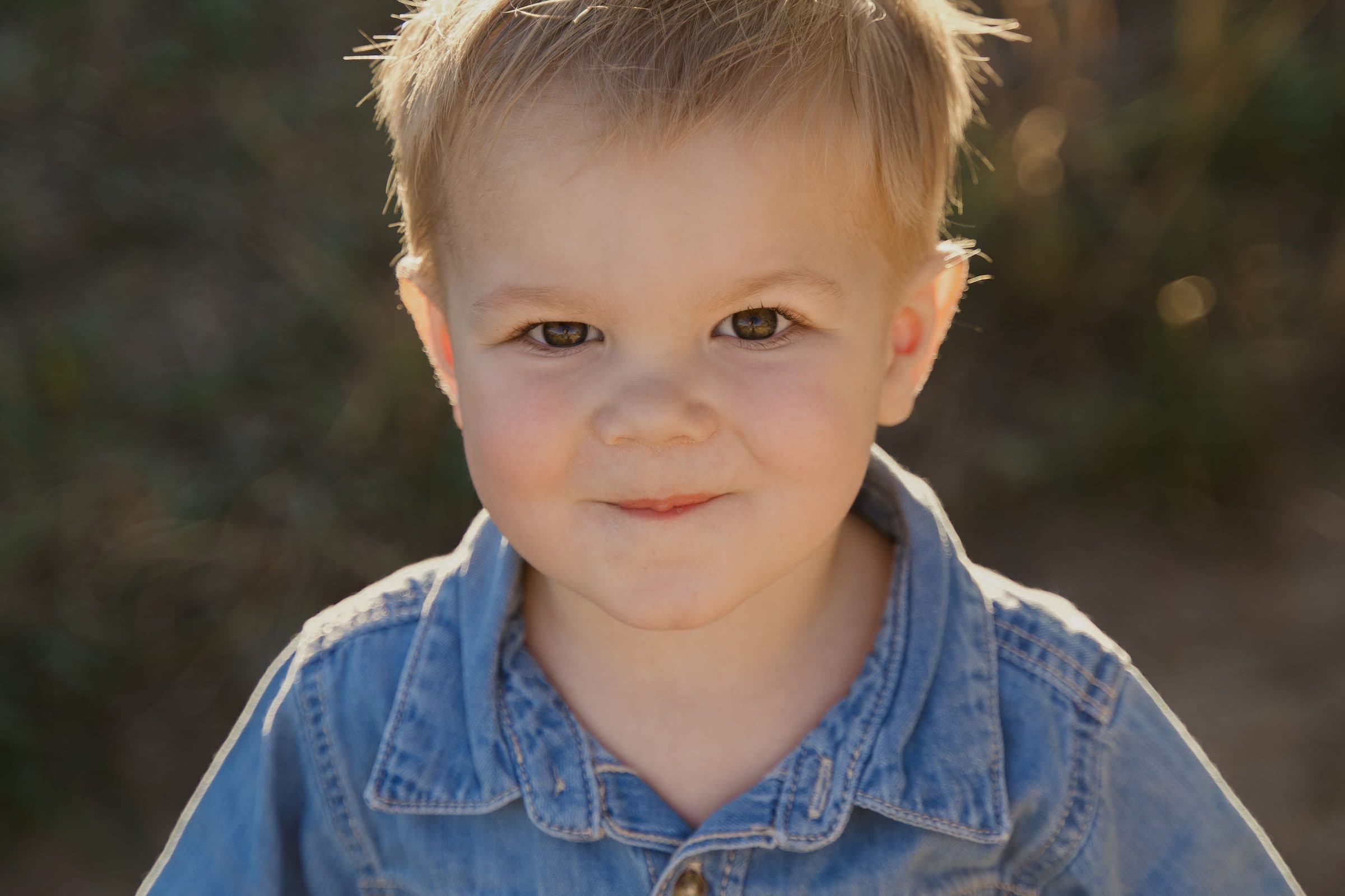 Close-up of a smiling toddler boy in a denim shirt, backlit by the sun