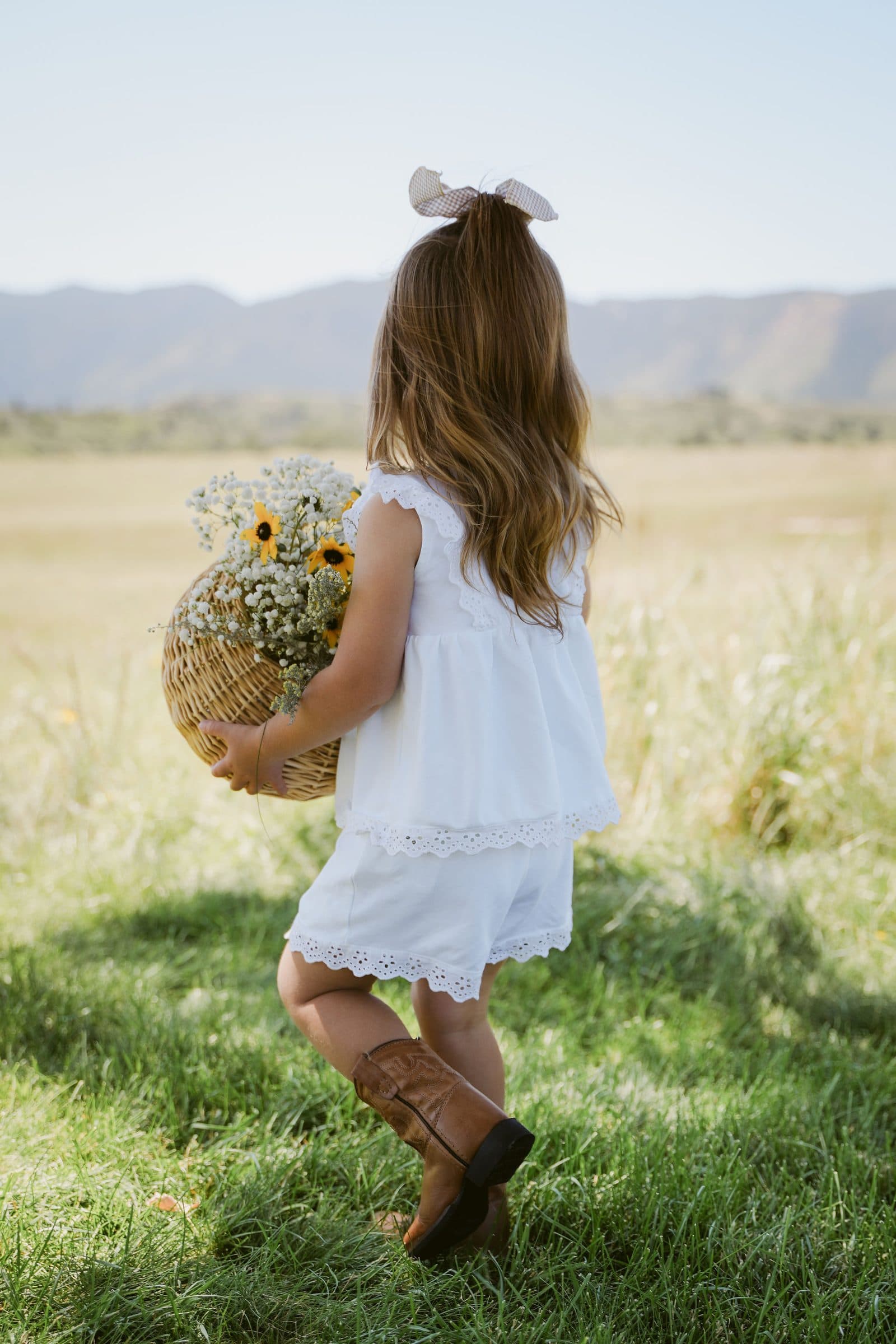 Little girl in a white dress and boots carrying a basket of sunflowers through a meadow