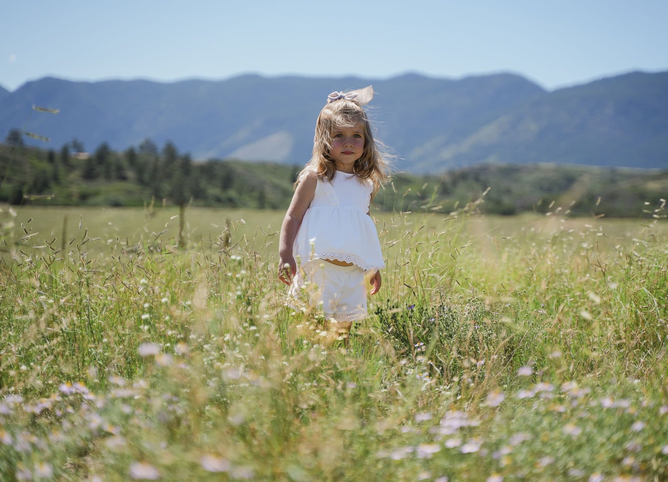 Little girl in white walking through wildflowers with mountains behind her