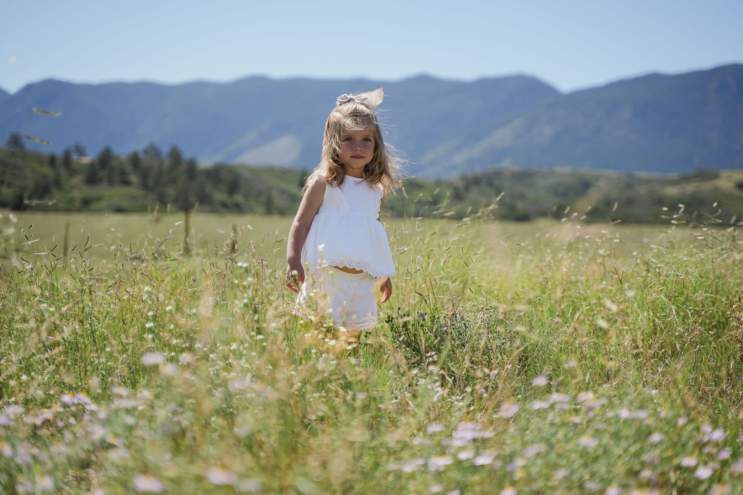Little girl in white walking through wildflowers with mountains behind her