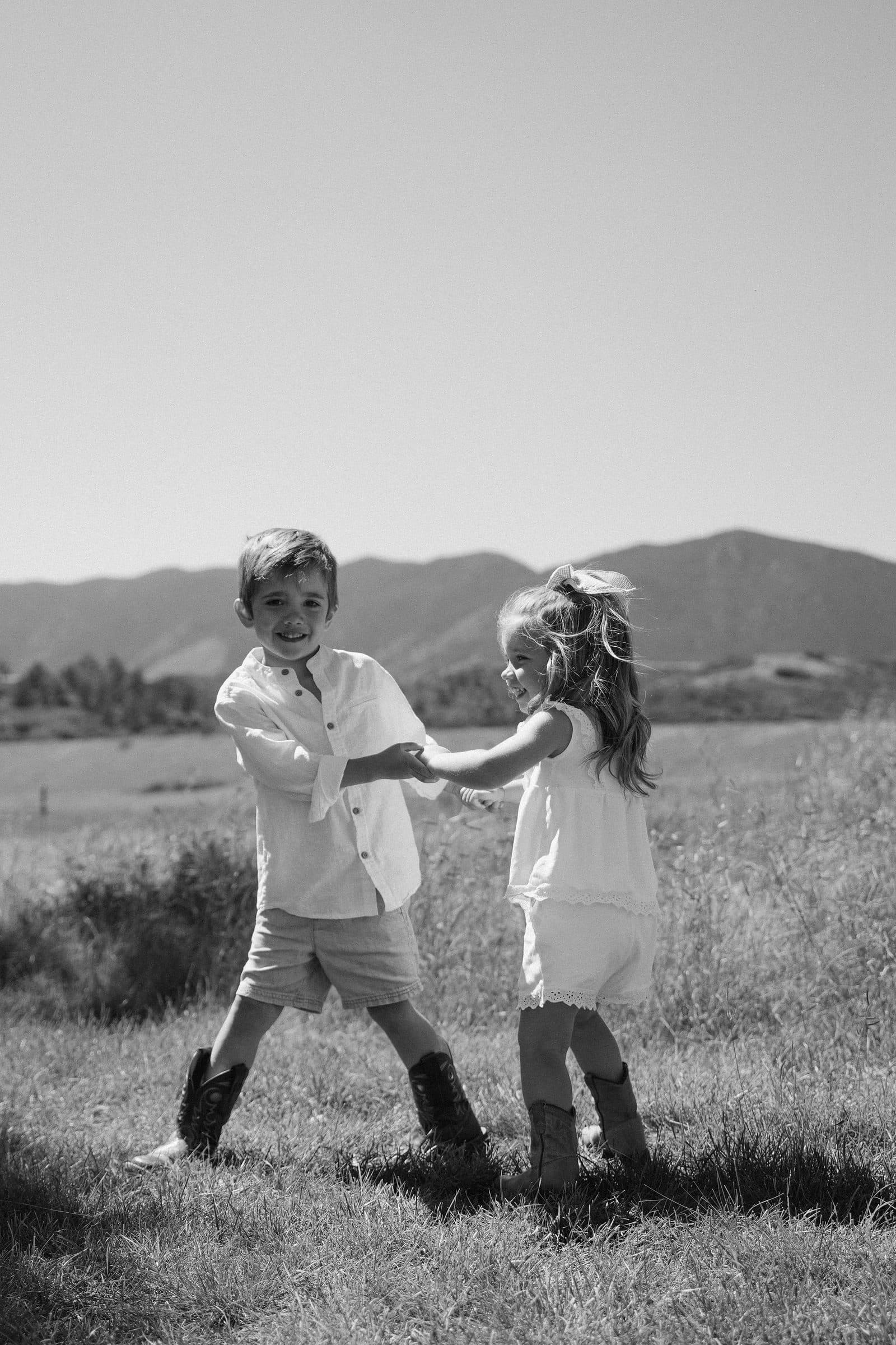 Black and white of a brother and sister holding hands and dancing in a field below the mountains
