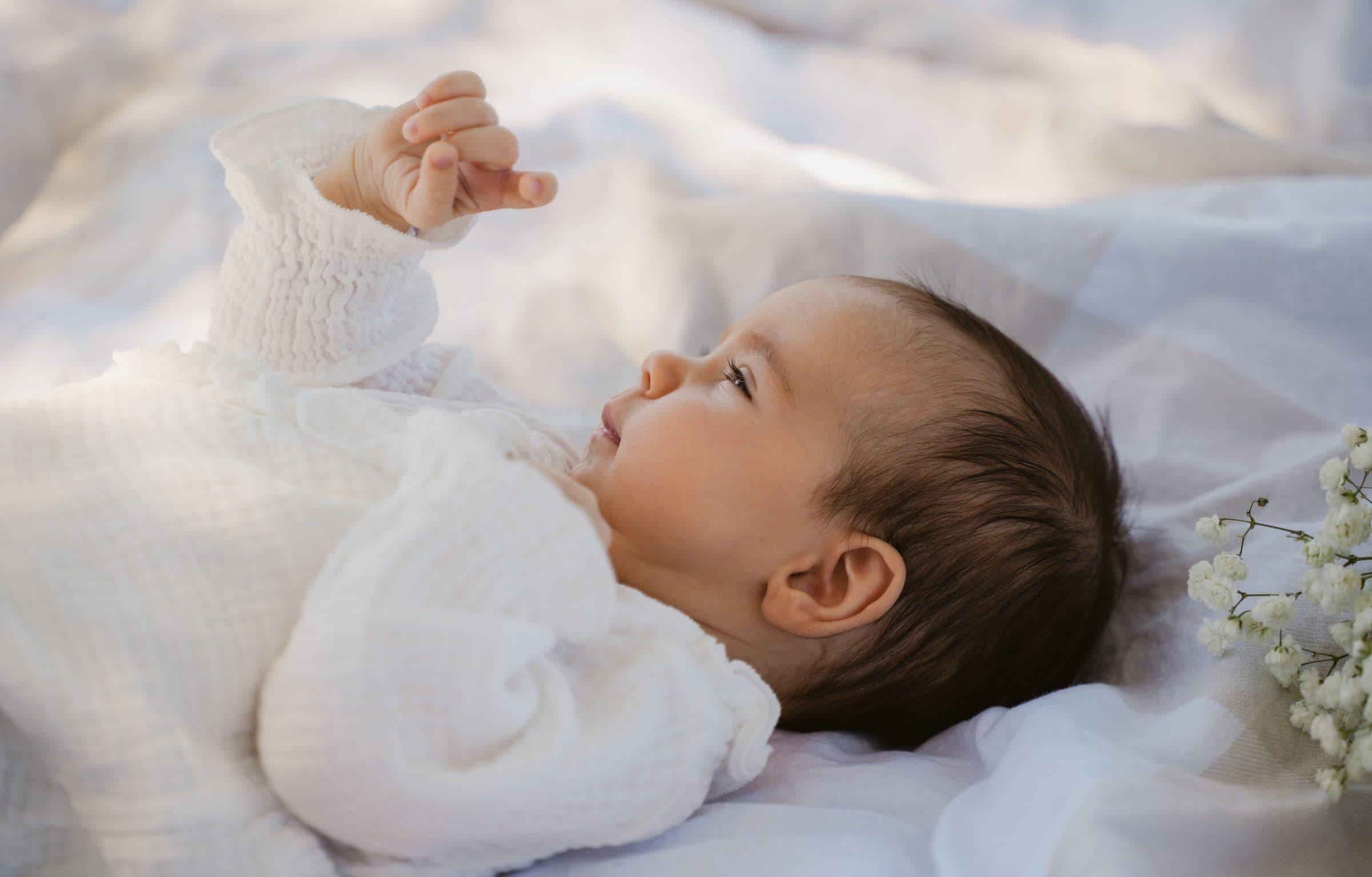 Newborn in a white knit outfit lying on soft linens, reaching one hand up