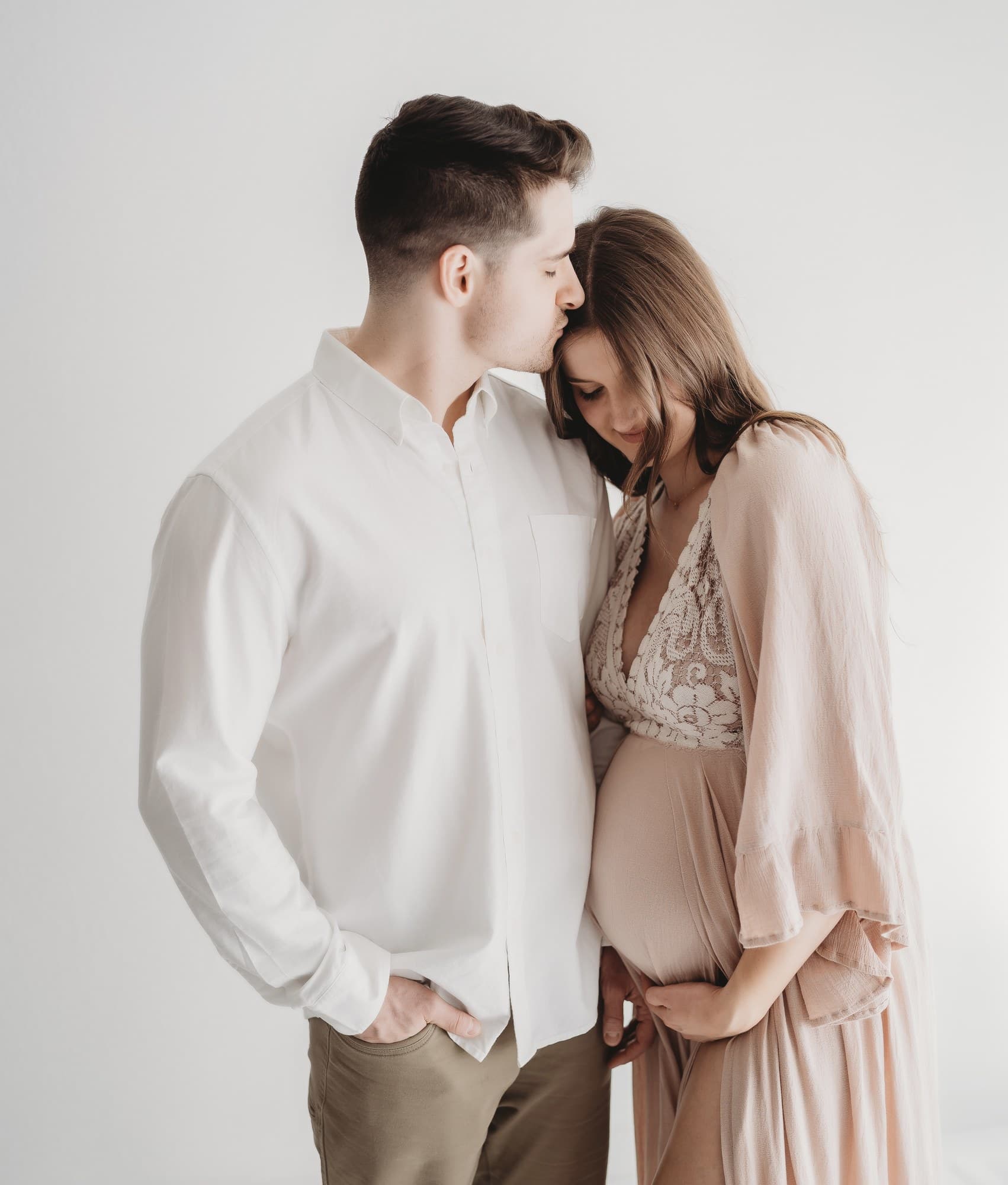 Expectant couple, forehead kiss, blush dress in a bright studio