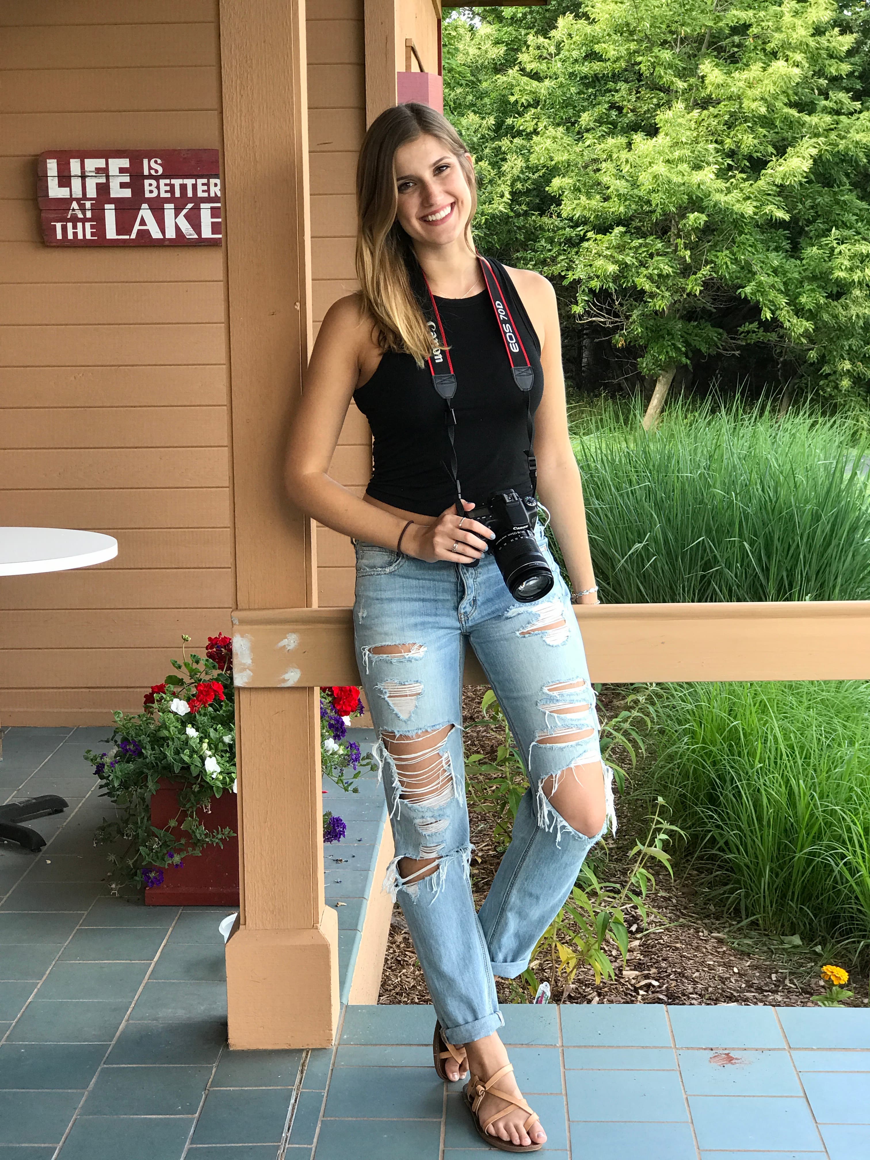 Olivia smiling on a lake-house porch, a camera hanging from a strap around her neck