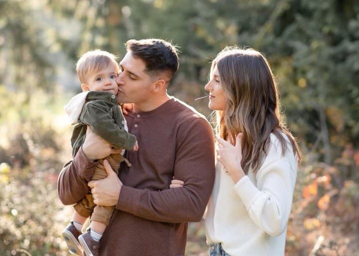 Father kissing a toddler on the cheek while mother looks on in a sunlit field
